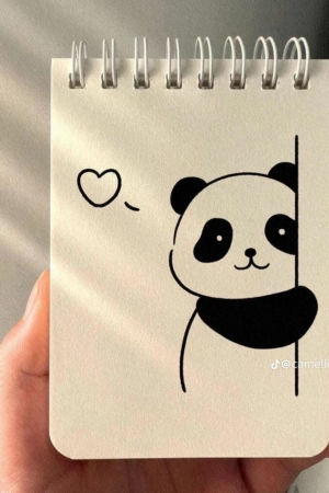 Uma mão segura um caderno espiral com um desenho fofo de um panda e um coração ao lado da cabeça, exibindo talentos incríveis em desenho.