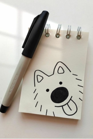 Um esboço de um cão brincalhão com a língua de fora, cuidadosamente desenhado em um pequeno bloco de notas em espiral ao lado de uma caneta. Ideal para aqueles que amam desenho e abraçam sua paixão por desenho.