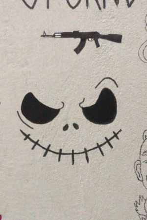 Para desenhar - Uma parede apresenta grafite de um smiley de boca costurada com órbitas oculares escuras, lembrando Jack Skellington, e uma silhueta preta de um rifle acima dele. Outros desenhos parciais são visíveis ao redor dele.