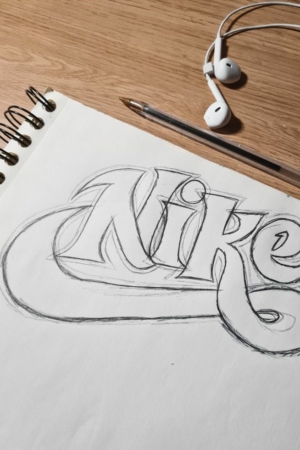 Para desenhar - Um caderno de esboços exibindo um logotipo "Nike" desenhado à mão em texto estilizado repousa sobre uma superfície de madeira ao lado de uma caneta esferográfica e um par de fones de ouvido brancos, mostrando o talento do artista para criar desenhos.