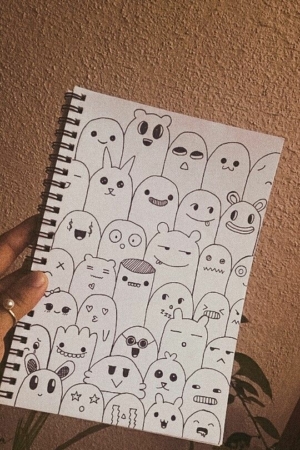 Caderno encadernado em espiral com personagens fofos desenhados à mão, semelhantes a fantasmas, segurados por uma mão, com fundo texturizado marrom. Para desenhar no caderno