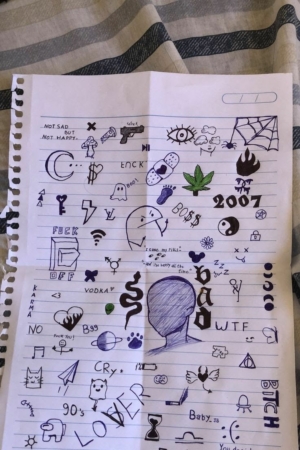 Página do caderno adornada com vários desenhos de criação, apresentando símbolos, palavras e um esboço de uma cabeça sem rosto no centro. Para Colorir.