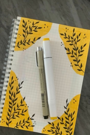 Um caderno com desenhos de folhas amarelas nos cantos, uma caneta de tinta cinza e um marcador branco e amarelo repousam sobre uma superfície escura. A capa do caderno adiciona um toque artístico aos seus itens essenciais de escrita.