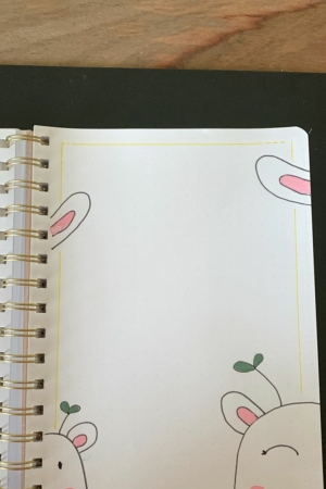 Caderno aberto com encadernação em espiral, com coelhinhos fofos e brotos de plantas em uma página pontilhada. A capa do caderno exibe desenhos encantadores desses elementos adoráveis, adicionando um toque de capricho às suas anotações.