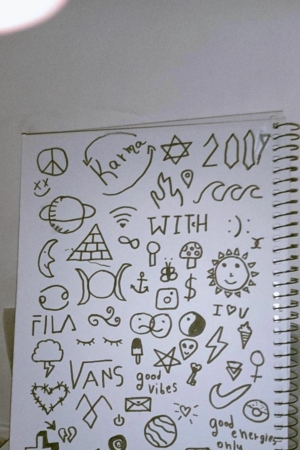 Uma página de caderno repleta de vários rabiscos, incluindo símbolos, estrelas, luas, nuvens e frases como "boas vibrações" e "COM". Para desenhar.