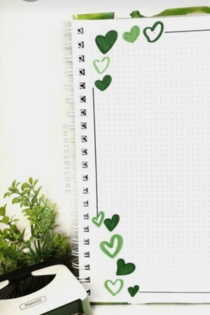 Caderno com uma página pontilhada, decorado com corações verdes ao longo da borda e um desenho intrincado na capa do caderno. Uma caneta e uma planta estão próximas.