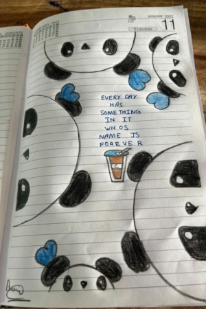 Agenda aberta com rostos de pandas desenhados à mão e corações azuis ao redor de uma citação que diz "Todo dia tem algo dentro cujo nome é para sempre". A capa do caderno apresenta um desenho intrincado, o que a torna um acessório charmoso para qualquer amante de papelaria.