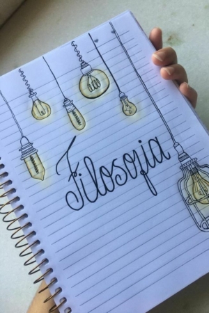 Uma mão segurando um caderno com "Filosofia" escrito na página pautada, decorado com desenhos de lâmpadas penduradas.