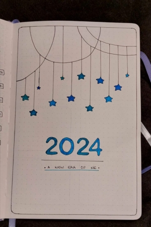 Uma capa de bullet journal para 2024, decorada com estrelas azuis penduradas e o texto "Uma Nova Era para Mim" na parte inferior.