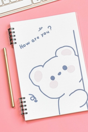 Um desenho fofo de um urso na capa do caderno com o texto "Como vai você?", ao lado de uma caneta dourada em uma superfície rosa.