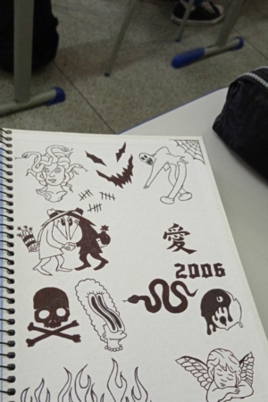 Um caderno com desenhos em tinta preta, representando caveiras, uma cobra, uma pessoa alada e o ano "2006" em uma página branca. Desenhos de criação dão um toque pessoal e fácil de fazer no caderno. Para Colorir.