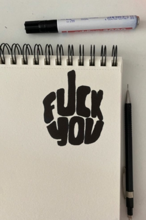 Caderno de desenho espiral com texto em negrito "FUCK YOU" em marcador preto; caneta preta e marcador ao lado, tornando-o um caderno fácil para fazer desenhos ou Desenhos de criação. Para Colorir.